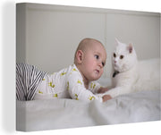 Canvas Schilderijen - Baby kijkend naar een witte kat op een bed - 90x60 cm - Wanddecoratie